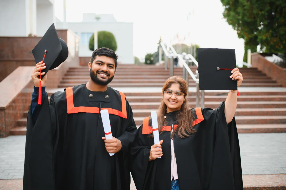 Two Indian graduates in gowns celebrating with caps and diplomas